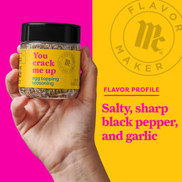 A hand holds a jar of Flavor Makers You Crack Me Up egg topping seasoning, with a yellow background showing the Flavor Maker logo and the flavor profile.