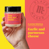 A hand holds a jar of Flavor Makers You Mac Me Smile mac and cheese seasoning with a red background that says Flavor Profile: Garlic and Parmesan Cheese.