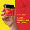 A hand holds a jar of Flavor Maker pizza topping seasoning with a red background that says Flavor Profile: Garlic, oregano, and red pepper.