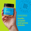A hand holds a jar of Flavor Makers Im So Hooked salmon topping seasoning, with a blue background that says Flavor Maker and the flavor profile of garlic, sesame, and green onions.