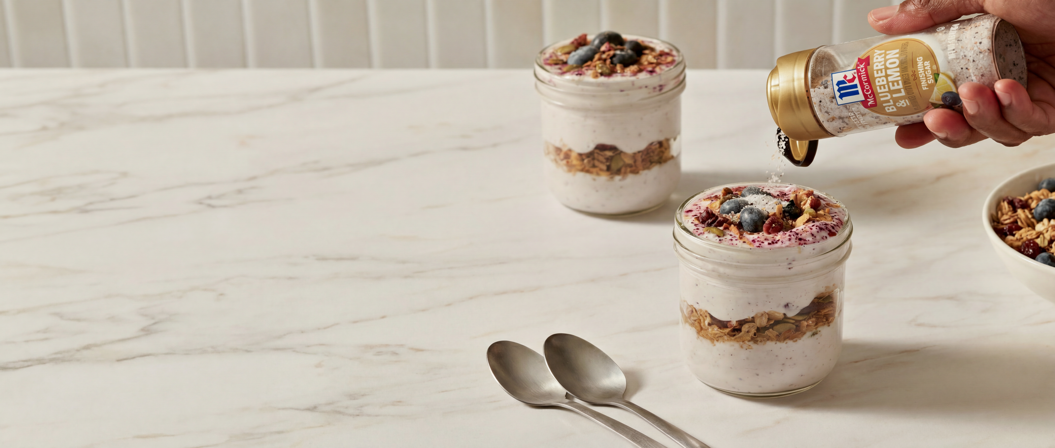 Person pouring finishing sugar into a jar of yogurt parfait on a marble surface