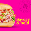 A Flavor Maker sandwich with lettuce, tomato, and salami on a white bun with the words Savory &amp; bold in yellow.