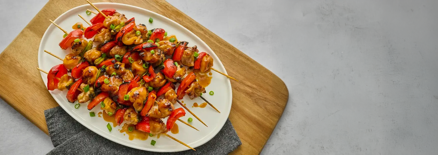 Grilled chicken and red bell pepper skewers on a white oval platter, placed on a wooden cutting board with a gray napkin on a light gray surface.
