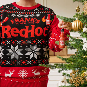 A person wearing a Frank s RedHot holiday sweater hangs a miniature bottle of Frank s RedHot Original Cayenne Pepper Sauce as an ornament on a Christmas tree.