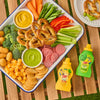 A colorful snack board featuring pretzels, vegetables, and cheese is arranged on a wooden table next to bottles of French's MINION Classic YELLOW MUSTARD and French's GOOMI'S GREEN MUSTARD, which are also shown as dips on the platter.