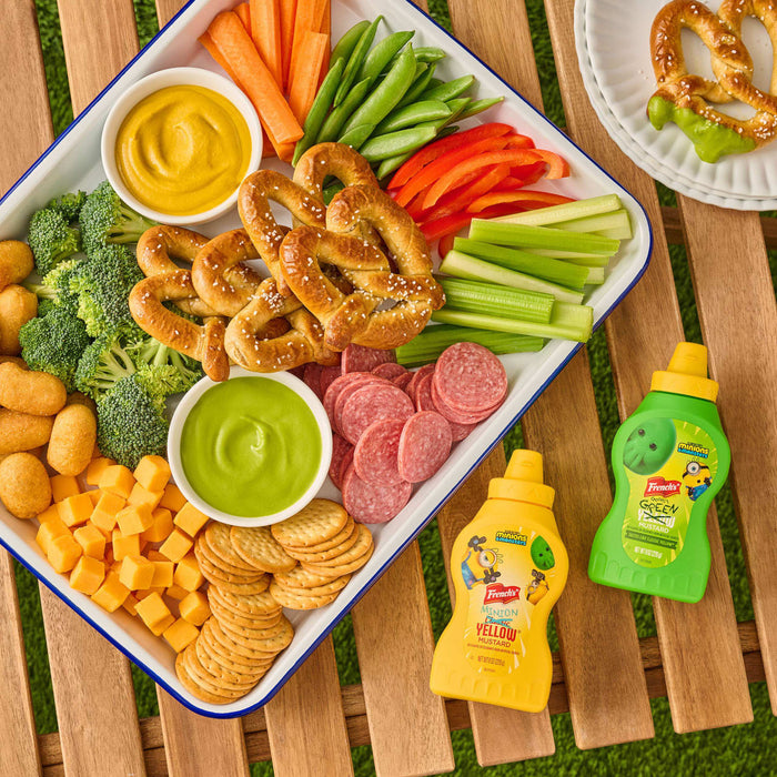 A colorful snack board featuring pretzels, vegetables, and cheese is arranged on a wooden table next to bottles of French's MINION Classic YELLOW MUSTARD and French's GOOMI'S GREEN MUSTARD, which are also shown as dips on the platter.