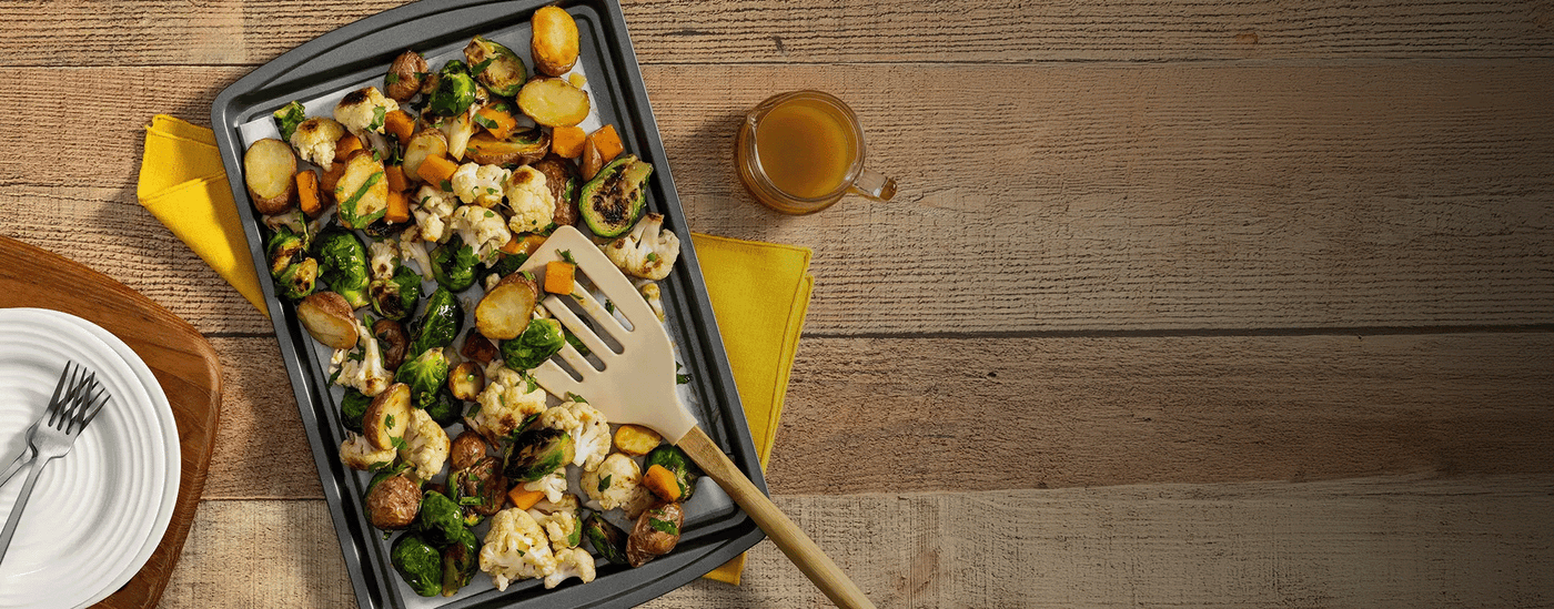 Roasted vegetables on a baking tray with a wooden spatula, on a wooden table with a glass of juice.