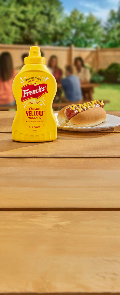 french's classic yellow mustard next to a hot dog with mustard on a picnic table with a family in the background