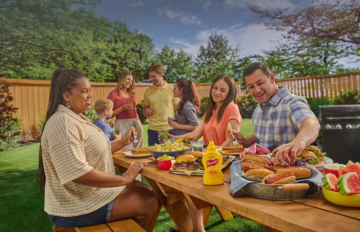 Family and friends enjoying a barbecue outdoors with food and drinks on a table.