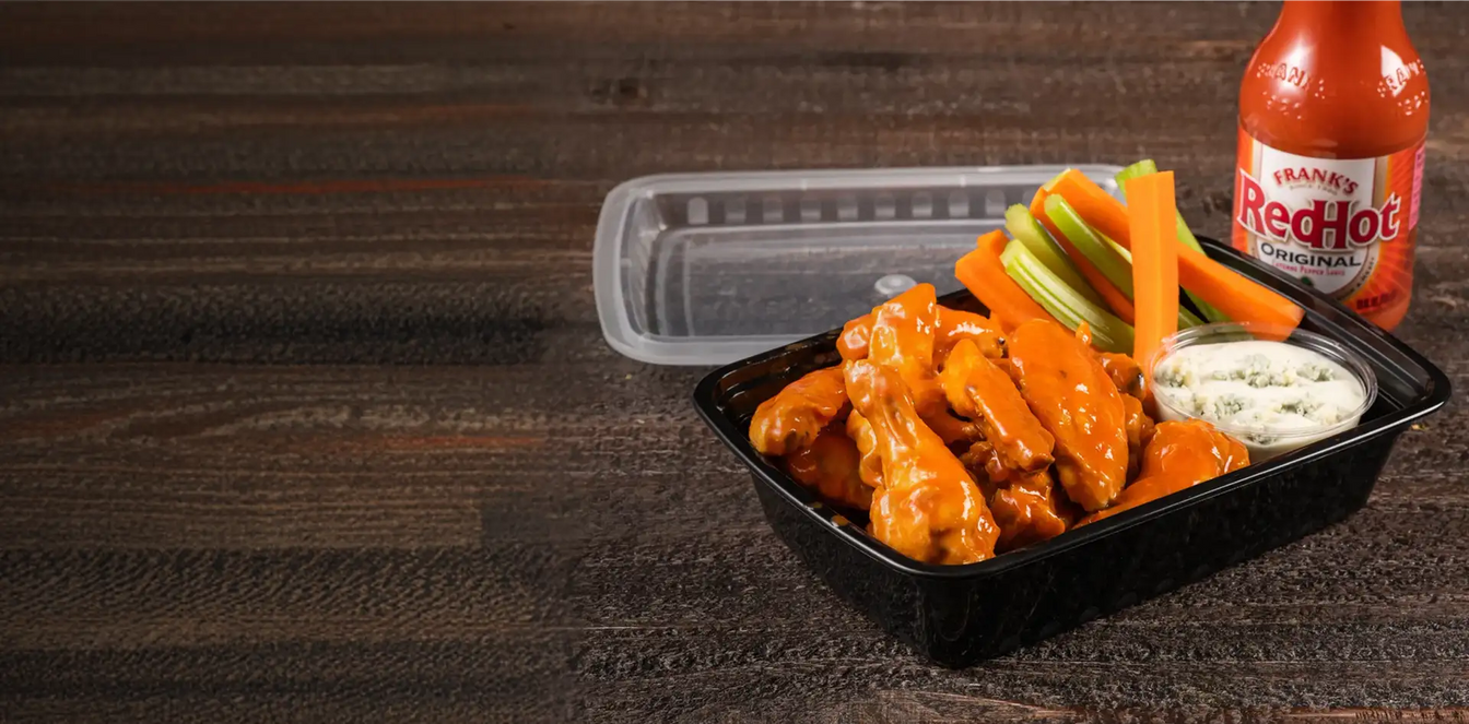 Platter of buffalo wings with celery, carrots, and blue cheese dressing next to a bottle of Frank's RedHot sauce on a wooden surface.