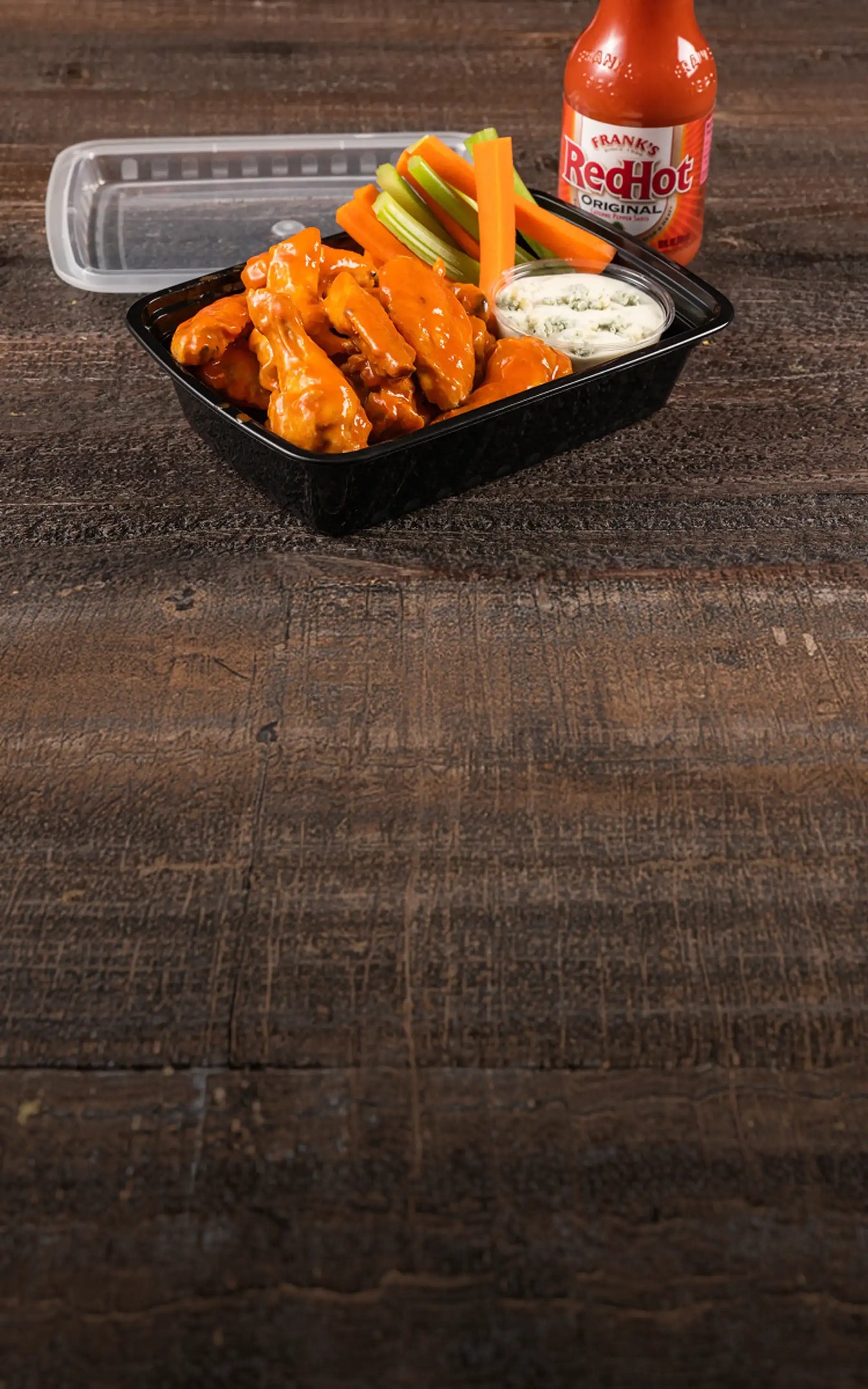 Black container of buffalo wings with celery, carrots, and ranch dressing on a wooden surface with a bottle of Frank's RedHot sauce.