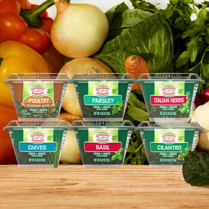 Six Gourmet Garden lightly dried herb and seasoning containers, including Gourmet Garden Basil (Case Of 72), are arranged on a wooden surface with fresh vegetables like peppers, onions, and cabbage in the background.