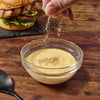 A hand sprinkles seasoning over a bowl of yellow sauce.