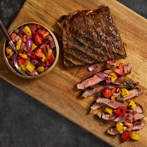 A grilled flank steak is presented on a wooden board, sliced and topped with a colorful tomato and pepper salsa.