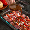 A person pours marinade over skewers of meat, onions, and red peppers in a glass baking dish.
