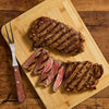 A grilled steak, partially sliced, is presented on a wooden cutting board alongside a carving fork, showcasing its juicy, medium-rare interior.