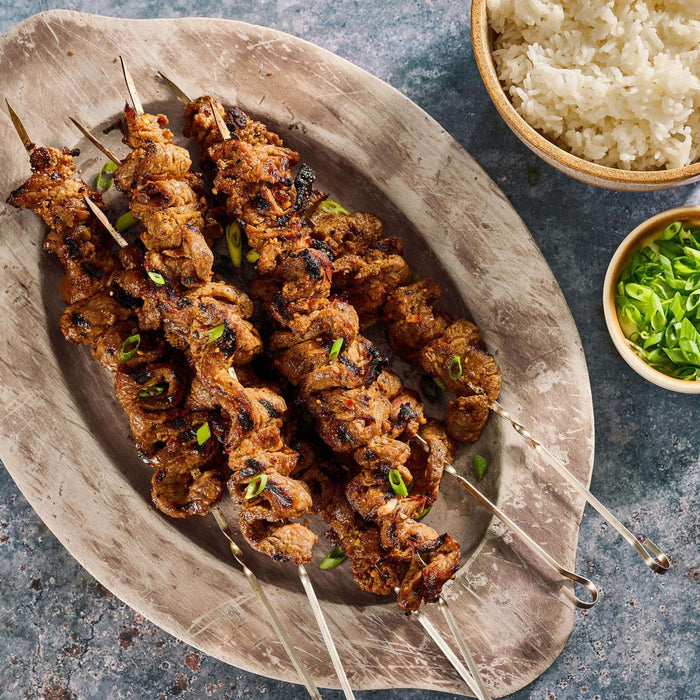 A platter of grilled meat skewers, prepared using Mccormick Grill Mates Asian Bbq Meat Marinade Mix 1.25 Oz, is served with a side of rice and chopped green onions.