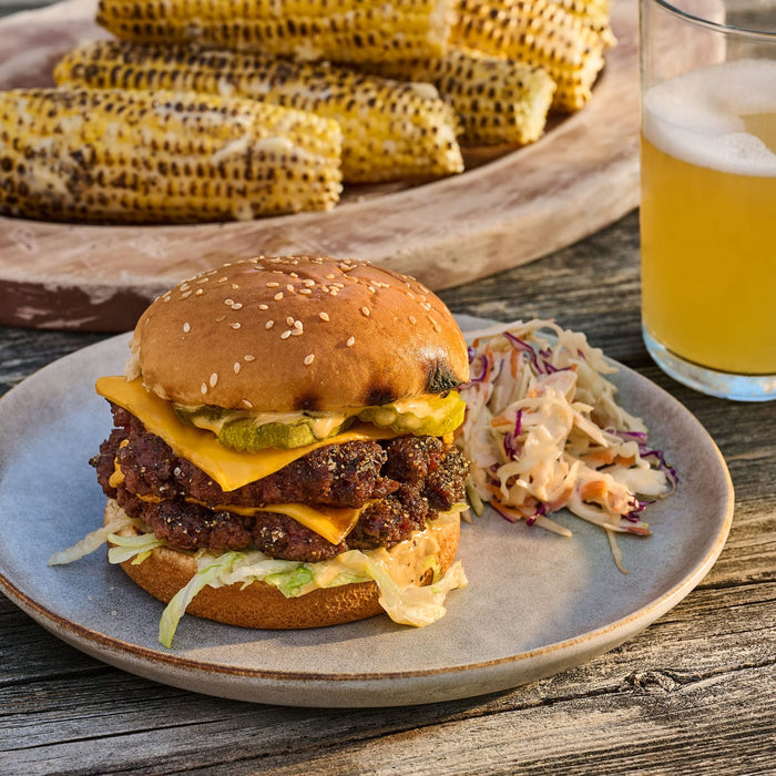 A juicy double cheeseburger, perfectly seasoned with Mccormick Grill Mates Signature Blend Seasoning, 6.53 Oz, is served on a rustic plate with coleslaw, with grilled corn and a beer in the background.