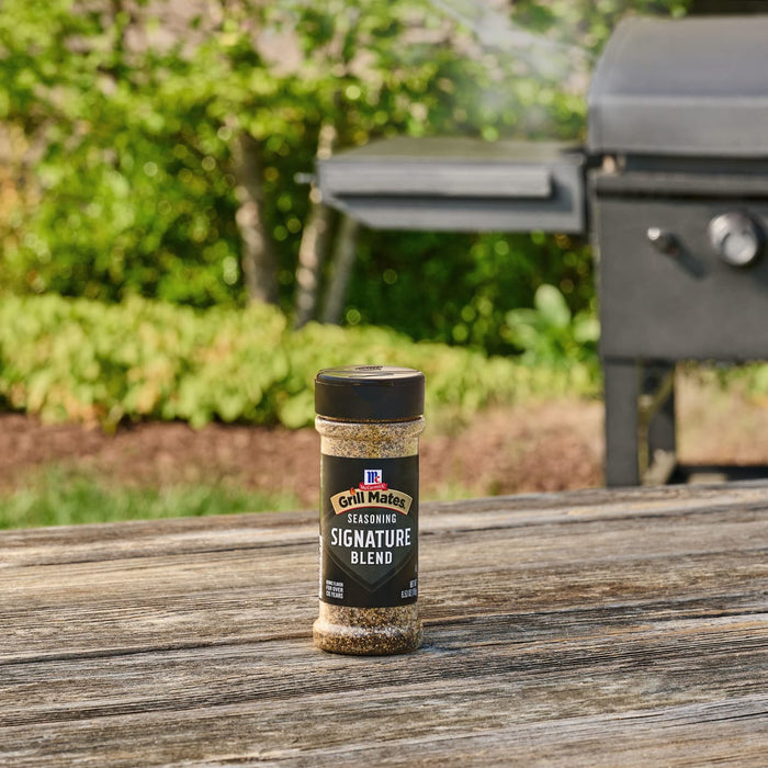 A bottle of Mccormick Grill Mates Signature Blend Seasoning, 6.53 Oz sits on a rustic wooden table with a barbecue grill ready for use in the sunny, green background.