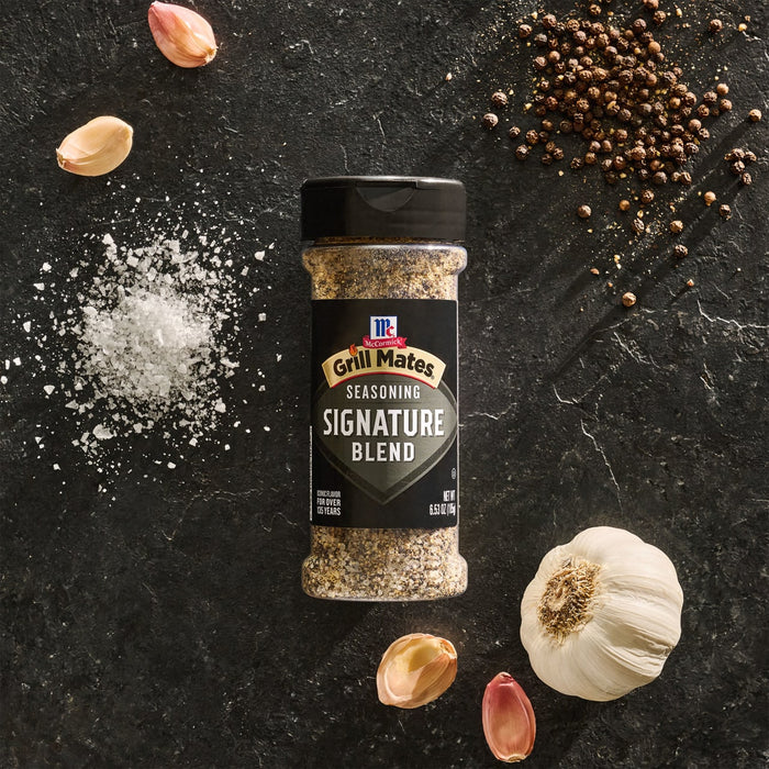 A bottle of Mccormick Grill Mates Signature Blend Seasoning, 6.53 Oz, is arranged on a dark slate surface, surrounded by its core ingredients of whole black peppercorns, coarse salt, and fresh garlic.