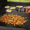 A hand sprinkles Mccormick Grill Mates Signature Blend Seasoning, 6.53 Oz over a pile of steaming breakfast potatoes and bacon cooking on a griddle next to several fried eggs.