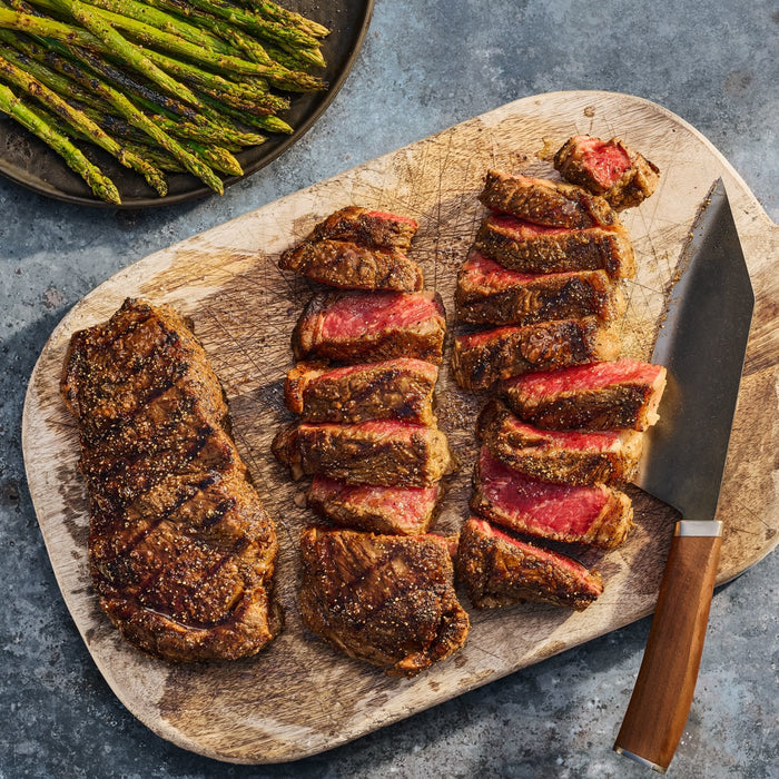 A perfectly grilled steak, seasoned with Mccormick Grill Mates Signature Blend Seasoning, 6.53 Oz, is sliced on a wooden board next to a side of roasted asparagus.