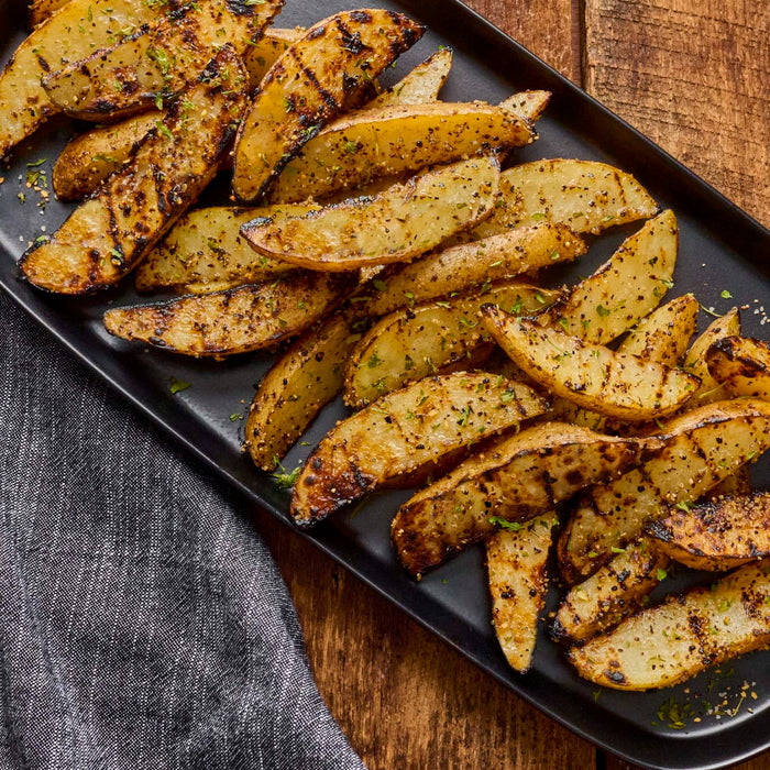 Grilled potato wedges seasoned with Mccormick Ppr Blk Crsh Gar Herb Sea Salt are arranged on a black platter, showcasing visible grill marks and herbs.