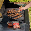 A person bastes meat skewers on a grill with a sauce made from a packet of Mccormick Grill Mates Asian Bbq Meat Marinade Mix 1.25 Oz, which is displayed next to a bowl of the marinade.