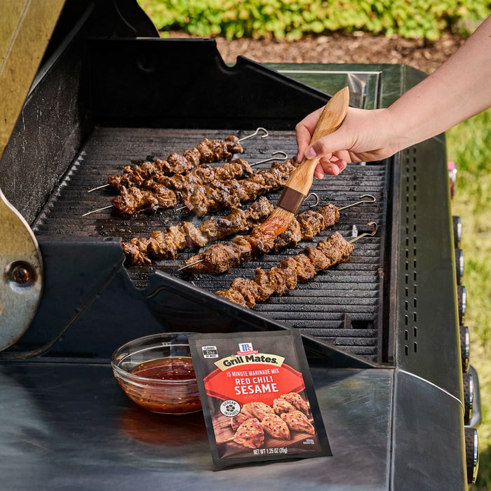 A person bastes meat skewers on a grill with a sauce made from a packet of Mccormick Grill Mates Asian Bbq Meat Marinade Mix 1.25 Oz, which is displayed next to a bowl of the marinade.