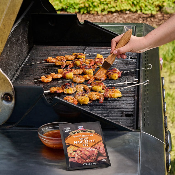 A person bastes shrimp and pineapple skewers on a grill, with a packet of Mccormick Grill Mates Hawaiian Style Bbq Meat Marinade Mix 1.25 Oz displayed in the foreground next to a bowl of the prepared marinade.