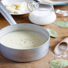 A pot of white sauce with pepper on top sits on a wooden table next to a pitcher of milk and a wooden spoon with flour.