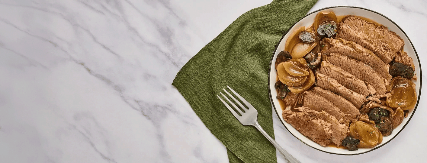A horizontal photo of a bowl of sliced meat with onions and mushrooms on a white surface, accompanied by a green napkin and a fork.