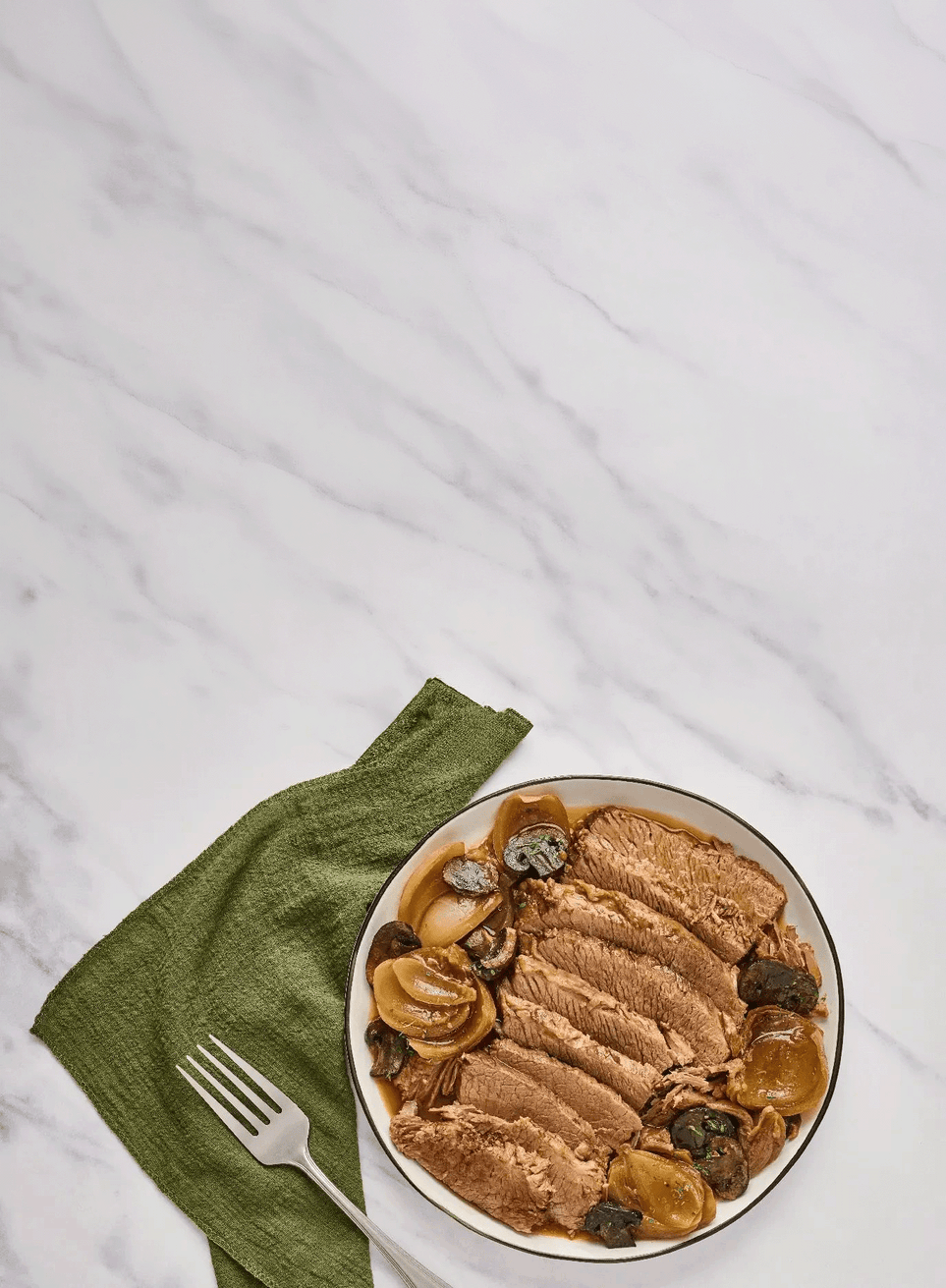Bowl of sliced meat with onions and mushrooms on a white surface with a green napkin and fork.