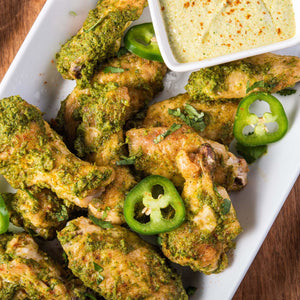 A plate of chicken wings covered in a green sauce with jalapeno slices and a side of white sauce.