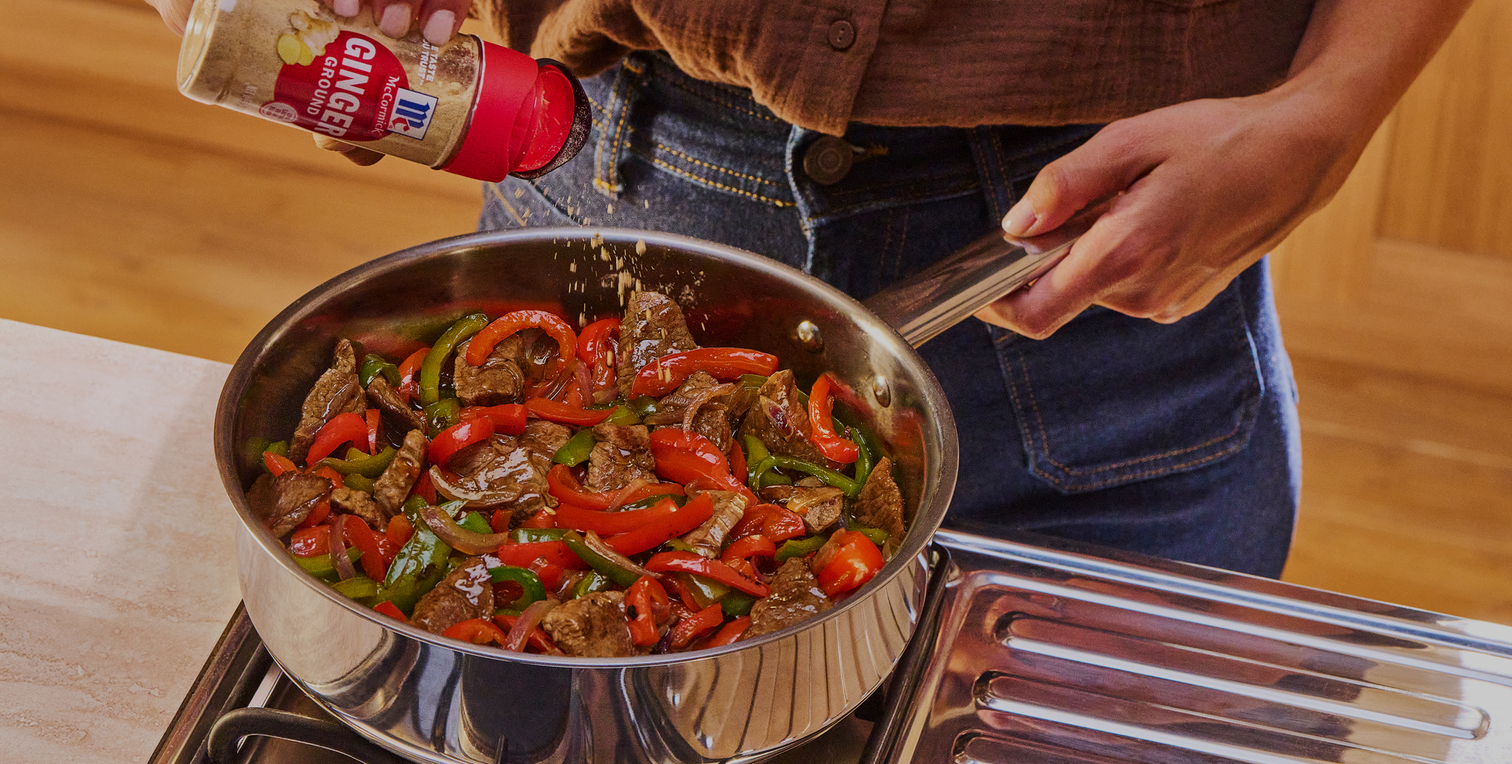 Person cooking a stir-fry with peppers and onions in a pan, using a bottle of sauce.