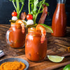Two loaded Bloody Mary cocktails in mason jars are garnished with celery, bacon, shrimp, and skewers of tomato and mozzarella, served alongside a small dish of red seasoning and a bottle of tomato juice.