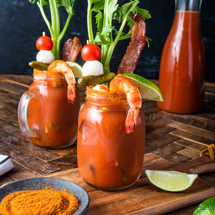 Two loaded Bloody Mary cocktails in mason jars are garnished with celery, bacon, shrimp, and skewers of tomato and mozzarella, served alongside a small dish of red seasoning and a bottle of tomato juice.