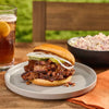 Brisket sandwich on a plate with a glass of iced tea and a bowl of potato salad in the background.