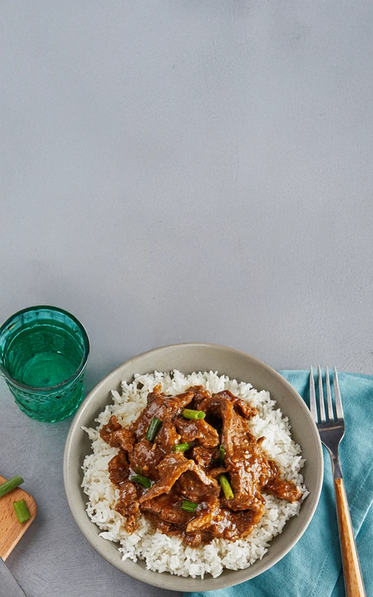 Bowl of rice with meat dish on a gray surface with a green glass and fork.