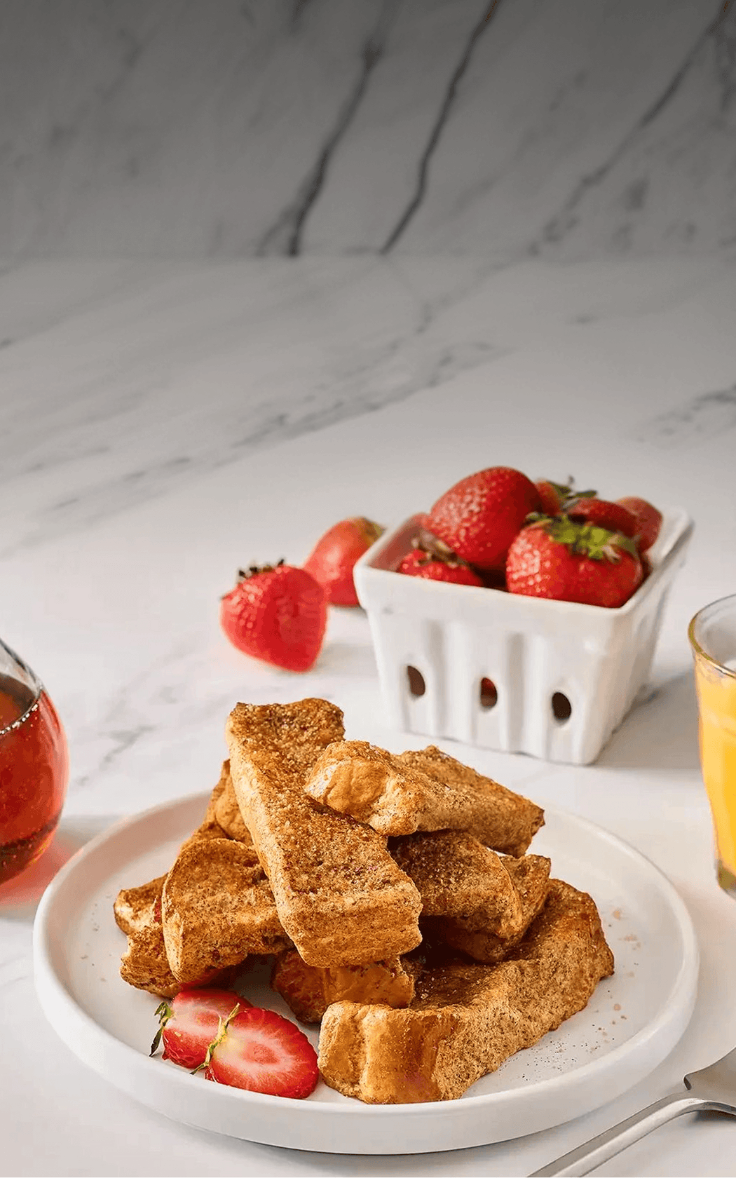 Stack of French toast on a plate with strawberries and a glass of orange juice on a marble surface.