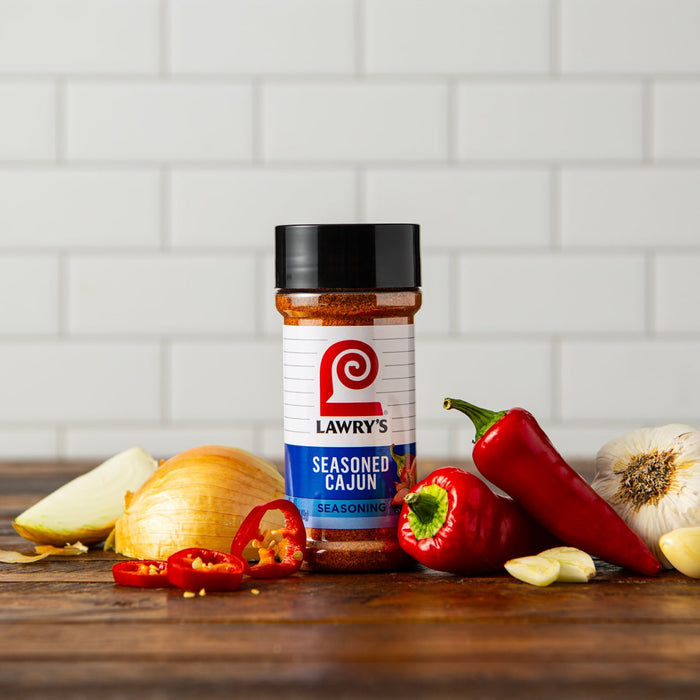 A bottle of Lawry's Seasoned Cajun seasoning sits on a wooden table with onions, garlic, and red peppers.