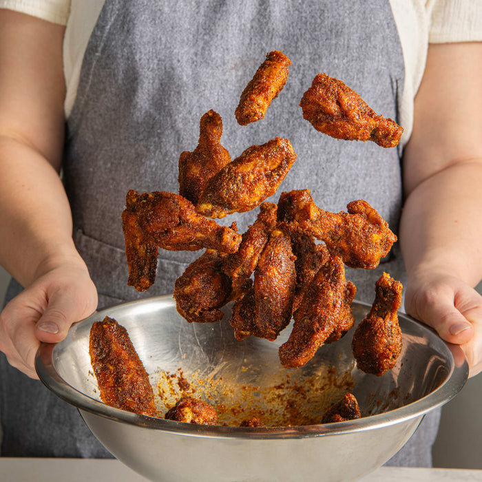Person holding a metal bowl with fried chicken wings, wearing a gray apron.