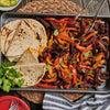 A tray of sizzling fajitas with tortillas, featuring strips of seasoned beef, sautACed onions, and red bell peppers, garnished with fresh cilantro.