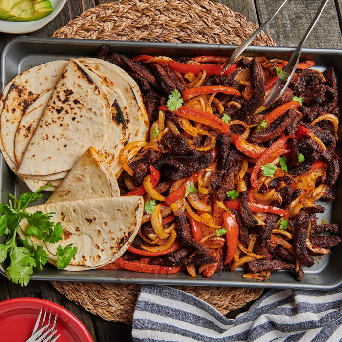 A tray of sizzling fajitas with tortillas, featuring strips of seasoned beef, sautACed onions, and red bell peppers, garnished with fresh cilantro.