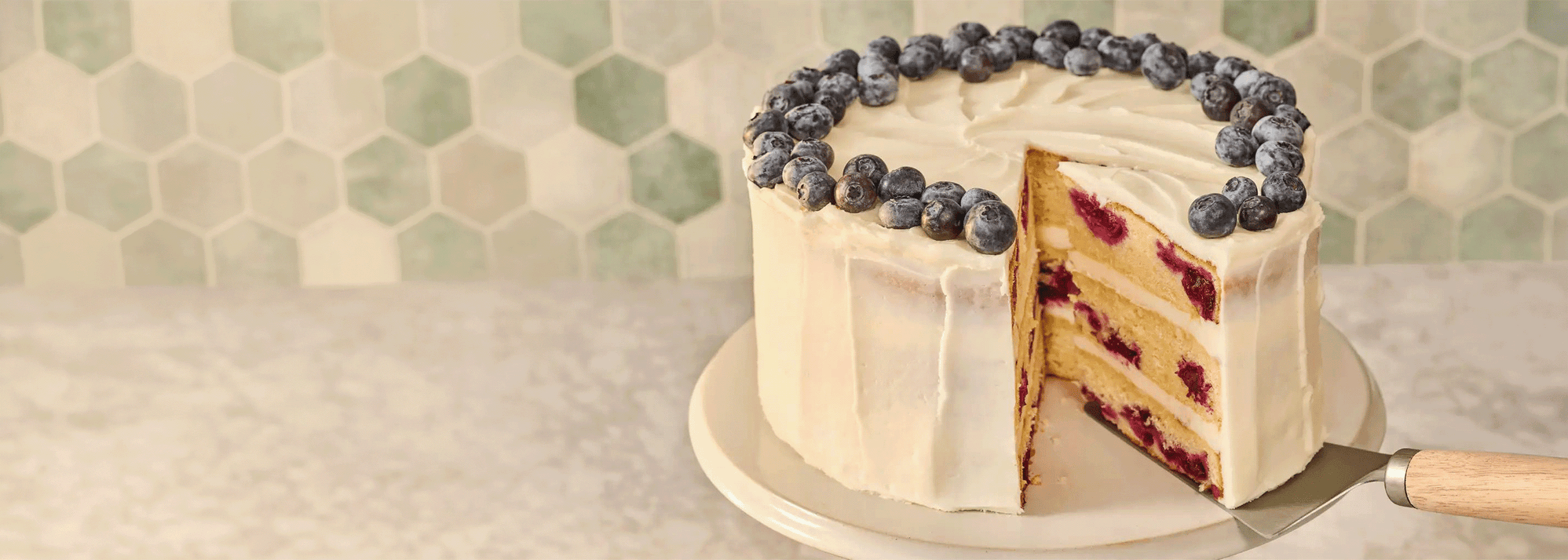 Horizontal image of a lemon blueberry cake with a hexagonal-patterned wall in the background