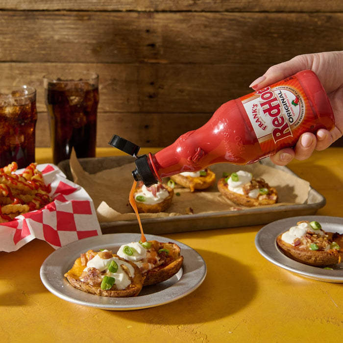 A hand pours Frank's RedHot Original Cayenne Pepper Sauce onto loaded potato skins topped with sour cream, bacon, and green onions.