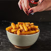 A person seasons a bowl of crispy golden fries with a McCormick Black Peppercorn Grinder over a dark marble surface.