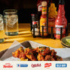 A tray of glazed chicken wings garnished with herbs sits on a table next to a condiment caddy featuring Frank s RedHot Original Cayenne Pepper Sauce, Cholula Hot Sauce, French s Classic Yellow Mustard, and McCormick Culinary salt and pepper grinders.