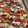 A close-up of a baking sheet with roasted vegetables, including red potatoes, zucchini, carrots, and red onions.