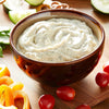 A bowl of creamy dip with green herbs sits on a wooden cutting board with sliced cucumbers peppers and tomatoes.
